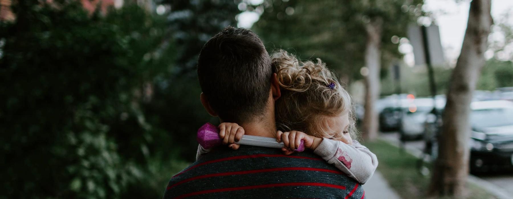 tired little girl rests her head on her father's shoulder as he carries her down the city sidewalk